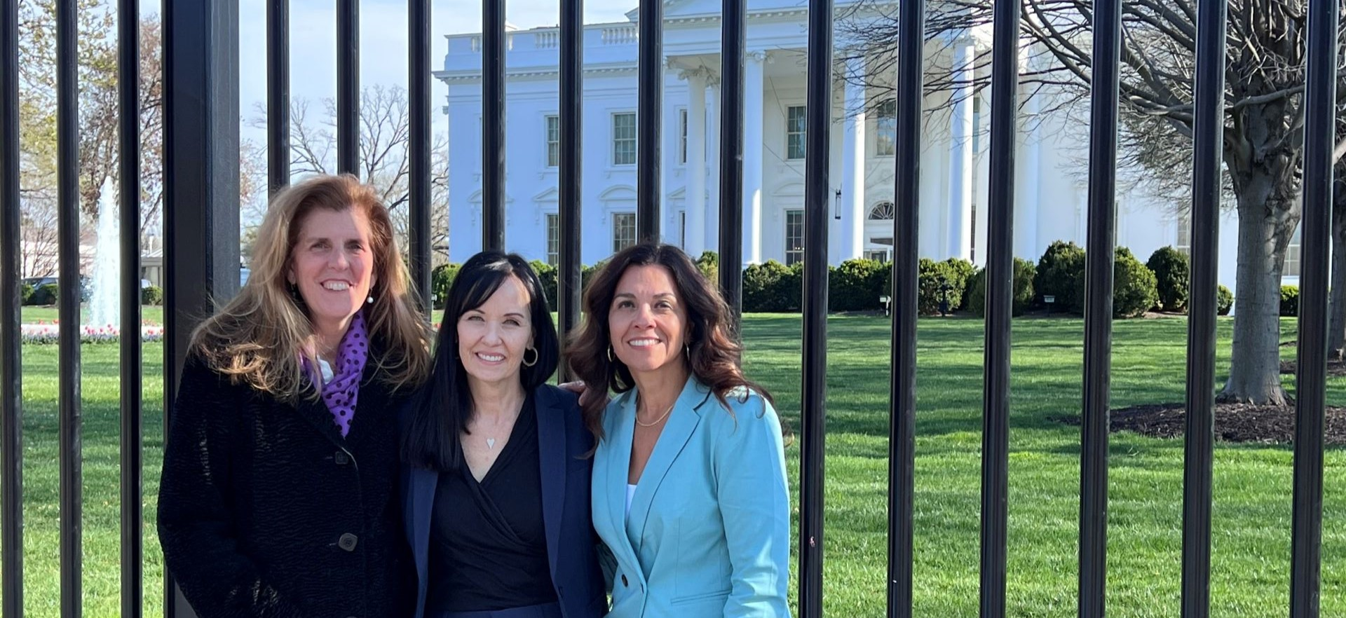 Stephanie Hoopes, Theresa Leamy, and Kiran Handa Gaudioso stand outside the gates of the White House and smile at the camera